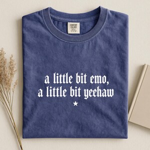 May include: A blue short-sleeved t-shirt with a crew neckline and a Comfort Colors label at the inner collar. The front features white text that reads, a little bit emo, a little bit yeehaw, centered above a small white five-pointed star graphic.