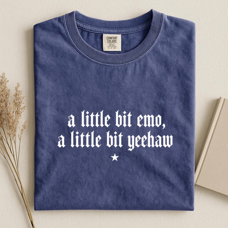 May include: A blue short-sleeved t-shirt with a crew neckline and a Comfort Colors label at the inner collar. The front features white text that reads, a little bit emo, a little bit yeehaw, centered above a small white five-pointed star graphic.