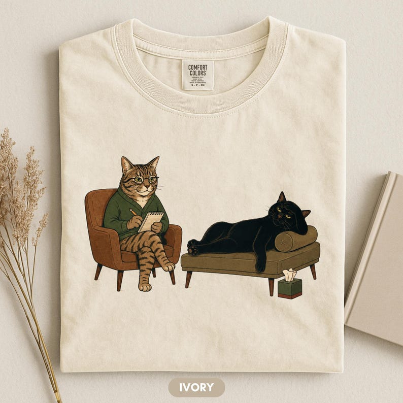 May include: An ivory t-shirt featuring a graphic of a tabby cat in a green sweater sitting in a brown armchair writing in a notebook, while a black cat lies on a brown chaise lounge next to a tissue box. The shirt has a Comfort Colors label.