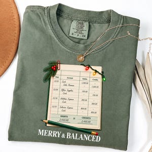 Può includere: Una t-shirt verde oliva con la grafica di una pagina di libro contabile decorata con una graffetta rossa, aghi di pino, bacche rosse e luci natalizie. Una matita verde è posizionata in basso. Il testo recita MERRY & BALANCED.