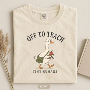 Peut inclure: Un t-shirt couleur ivoire avec une illustration d'une oie blanche portant des lunettes, une sacoche verte et une pile de livres surmontée d'une pomme rouge. Le texte "OFF TO TEACH" est inscrit au-dessus et "TINY HUMANS" en dessous.