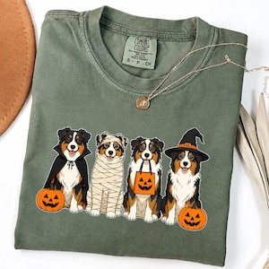 May include: An olive green t-shirt featuring a graphic of four Australian Shepherd dogs dressed for Halloween. One dog wears a vampire cape, one is wrapped as a mummy, one holds a jack-o'-lantern treat bag, and one wears a witch hat with a pumpkin.