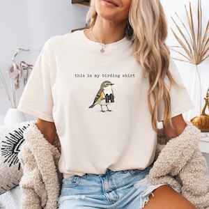 May include: A cream-colored short-sleeve t-shirt featuring a graphic of a small bird holding black binoculars. Above the bird, the text reads "this is my birding shirt" in a simple, black, typewriter-style font. The shirt has a crew neckline.