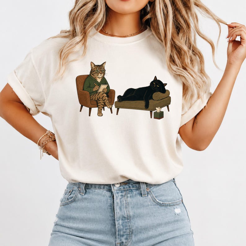 May include: A cream-colored t-shirt featuring a graphic of a tabby cat in a green sweater sitting in a brown armchair writing in a notebook, while a black cat lies on a green ottoman next to a small box of tissues on the floor.