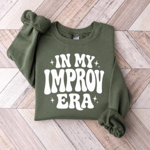 May include: An olive green crewneck sweatshirt featuring white text that reads IN MY IMPROV ERA in a bold, rounded font. The design is accented with small four-pointed star graphics positioned above and below the text on the front of the garment.