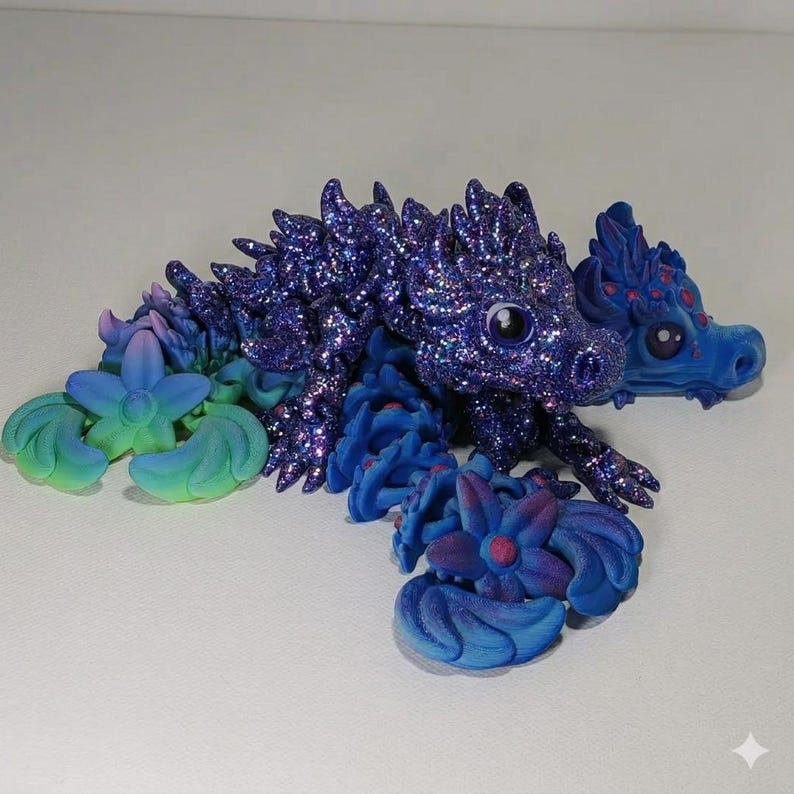 May include: A handcrafted fantasy sculpture of a dragon with a glittery, dark blue body and a lighter blue head. The dragon is adorned with blue flowers and leaves, with accents of pink and green.
