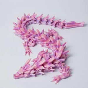 May include: A 3D-printed dragon figurine in shades of pink and purple. The articulated dragon has a detailed, spiky design, with a long, curving body and a head with pointed features. The dragon is posed on a white surface.