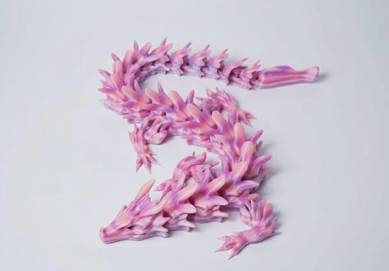 May include: A 3D-printed dragon figurine in shades of pink and purple. The articulated dragon has a detailed, spiky design, with a long, curving body and a head with pointed features. The dragon is posed on a white surface.