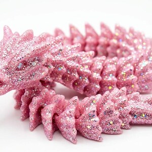 May include: A pink, glitter-covered dragon figurine. The dragon is curled, with a detailed head and spiky back. The pink color is accented with iridescent glitter, giving it a sparkling appearance. The figurine is on a white background.