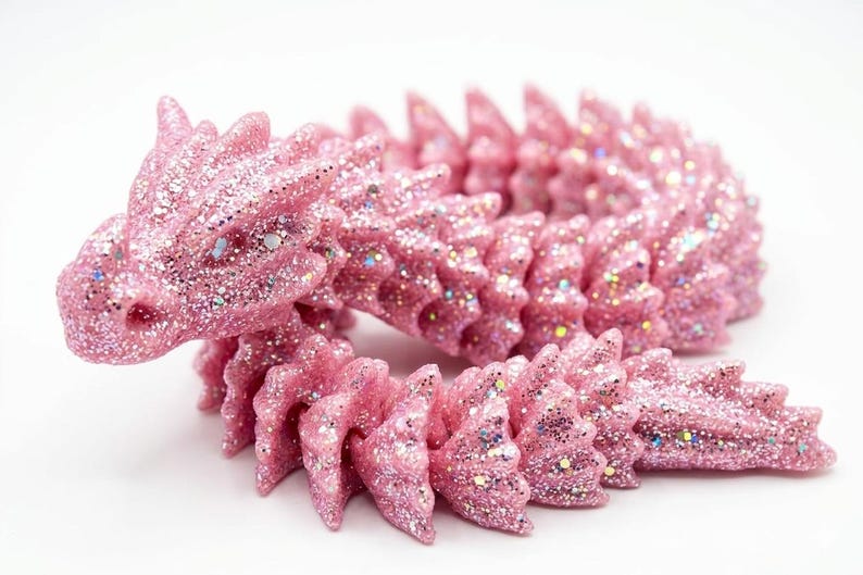May include: A pink, glitter-covered dragon figurine. The dragon is curled, with a detailed head and spiky back. The pink color is accented with iridescent glitter, giving it a sparkling appearance. The figurine is on a white background.
