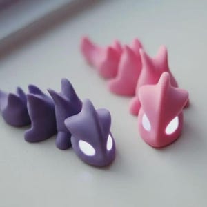 May include: Two small, stylized dragon figurines. One is purple, the other is pink. Both have a segmented body and a head with glowing white eyes. The figurines are on a light-colored surface.