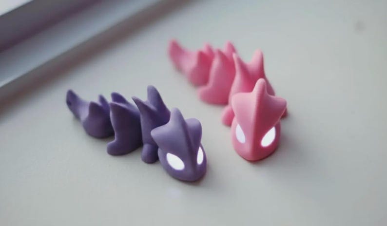 May include: Two small, stylized dragon figurines. One is purple, the other is pink. Both have a segmented body and a head with glowing white eyes. The figurines are on a light-colored surface.
