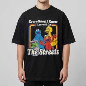 以下が含まれることがあります： 黒いTシャツ。セサミストリートのキャラクター、クッキーモンスター、エルモ、ビッグバード、オスカーのカラフルなグラフィック。テキスト「Everything I Know I Learned On The Streets」がキャラクターの上に表示されています。