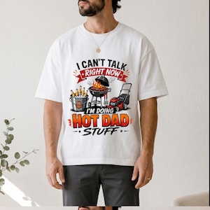 May include: White t-shirt with a graphic that reads "I CAN'T TALK RIGHT NOW I'M DOING HOT DAD STUFF." The graphic features a barbecue grill, a lawnmower, and a bucket of beer. The shirt is worn by a person.