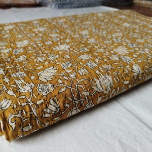 May include: A folded mustard yellow quilt featuring a repeating white floral pattern and visible white running stitch quilting throughout the fabric. The textile has a textured appearance with a dense arrangement of flowers and leaves across the surface.