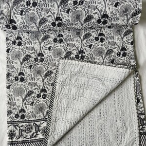 May include: A black and white quilt featuring a dense floral pattern with large blossoms and leafy vines. The fabric displays intricate hand-stitched kantha quilting lines throughout, with a striped border detail along the bottom edge.