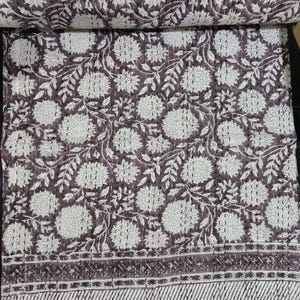Puede incluir: Una colcha de algodón en color marrón oscuro y blanco con un patrón floral repetitivo de grandes flores y hojas. La tela presenta bordados visibles de punto hilván por toda la superficie y el borde inferior tiene un acabado a rayas.