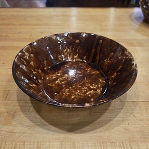 Puede incluir: Un cuenco redondo y poco profundo con un acabado brillante, que presenta un color base marrón oscuro con motas de color beige y crema más claras por toda la superficie interior y exterior, creando un aspecto texturizado y orgánico.