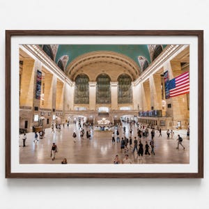 May include: A framed photograph of the Grand Central Terminal main concourse, featuring high arched windows, a teal ceiling, stone columns, and a large American flag hanging on the right side, with numerous people walking across the floor.