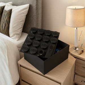 May include: A black storage box designed to look like a LEGO brick, with a hinged lid featuring LEGO studs. Inside the box are a remote control and a charging cable. The box is on a wooden bedside table next to a bed.