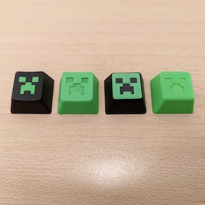 May include: Four square keyboard keycaps featuring a pixelated green face on a black or green background. The keycaps are arranged in a row and are likely for a computer keyboard.