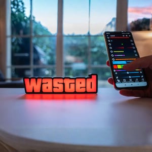 Op de afbeelding: Een rechthoekig LED-licht met het woord "WASTED" in rode blokletters. Het licht staat op een wit oppervlak. Een smartphone op de achtergrond toont een bedieningspaneel met kleuropties.