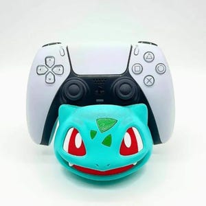 May include: A white and black PlayStation 5 controller with a teal Bulbasaur-shaped accessory. The Bulbasaur has red eyes and a green leaf design on its head. The controller has a black center section with white sides.