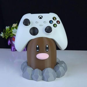 May include: A brown and grey 3D-printed Diglett-themed controller holder with a white Xbox controller on top. The holder has a pink nose and grey rock accents. The controller has coloured buttons.