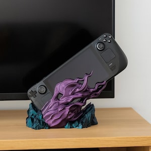 May include: A black handheld gaming console rests in a purple and teal decorative stand. The stand has a stylized, organic design. The console is angled, showcasing its buttons and screen. The stand is on a wooden surface.