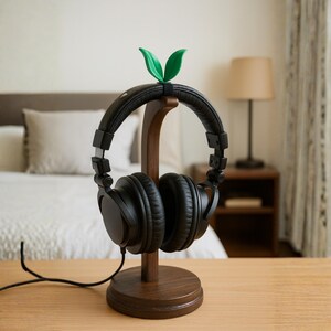 Op de afbeelding: Een donkerbruine houten hoofdtelefoonstandaard met zwarte over-ear hoofdtelefoon. De standaard heeft een groen blad detail aan de bovenkant. De standaard staat op een lichtbruin houten oppervlak.