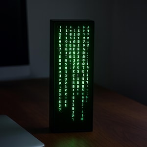 Puede incluir: Un reloj digital rectangular negro que muestra caracteres verdes en formato vertical. La pantalla muestra una serie de números y símbolos, que se asemejan a código. El reloj está sobre una superficie de madera, con un fondo oscuro.