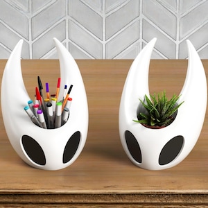 Pode incluir: Dois organizadores de mesa brancos com formato abstrato. Um contém canetas e marcadores coloridos, o outro uma pequena planta suculenta verde. Cada um tem três recortes ovais pretos e um topo curvo, sobre uma superfície de madeira.