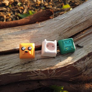 May include: Three colorful, square-shaped keycaps featuring cartoon character designs. The keycaps are orange, white, and green, with detailed facial features. They are resting on a piece of weathered wood.