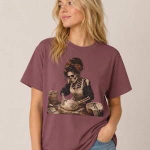Op de afbeelding: Een mauve t-shirt met korte mouwen en een grafische print van een skelet met een schort en een hoofddoek met patroon, dat deeg kneedt op een bebloemd oppervlak naast een zak met het opschrift FLOUR en een gevlochten mand.