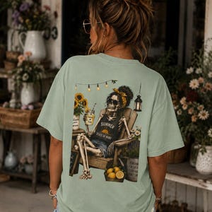 May include: A sage green short-sleeve t-shirt featuring a graphic on the back of a skeleton sitting in a wooden chair. The skeleton wears a yellow headband and sunglasses, holding a drink. Text reads "SUMMER STATE OF MIND" with sunflowers.