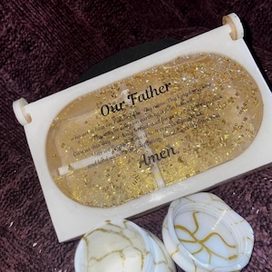 Puede incluir: Una pieza decorativa de resina con el texto "Padre Nuestro" y la oración del Señor. La pieza es rectangular con bordes redondeados, llena de purpurina dorada y tiene un marco blanco. Se ven dos posavasos a juego.