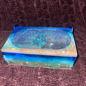 Puede incluir: Caja rectangular de resina con un diseño de temática oceánica azul y blanco. La caja presenta una tapa transparente con purpurina azul y pequeñas piedras, y los lados están decorados con capas de resina azul y blanca, que se asemejan a olas y arena.