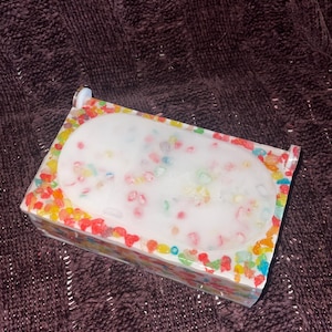 Puede incluir: Una pastilla de jabón rectangular con un centro ovalado blanco y adornos coloridos en forma de caramelos. El jabón está encerrado en un recipiente rectangular transparente, con los adornos coloridos en los bordes. El fondo es una tela oscura y texturizada.