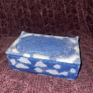 Puede incluir: Una caja rectangular de resina azul con tapa, decorada con formas de nubes blancas. La caja tiene un aspecto brillante y texturizado. El fondo es una tela oscura y texturizada.