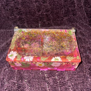 Puede incluir: Una caja rectangular de resina rosa con tapa transparente. La caja está decorada con flores blancas y hojas verdes. La tapa está llena de purpurina dorada y detalles en rosa y dorado. La caja tiene un asa transparente a cada lado.