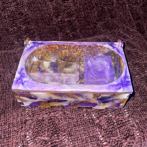 Puede incluir: Una caja rectangular de resina con tapa transparente, con un diseño veteado de purpurina morada, blanca y dorada. La caja tiene un pequeño compartimento en el interior y está sobre un fondo morado oscuro.
