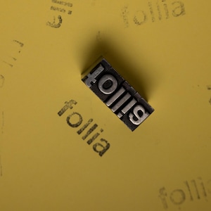 Op de afbeelding: Een metalen letterpress-blok met het woord "follia" in een schreeflettertype. Het blok ligt diagonaal op een gele ondergrond, met het woord "follia" vaag afgedrukt. De afbeelding is goed belicht en toont details.