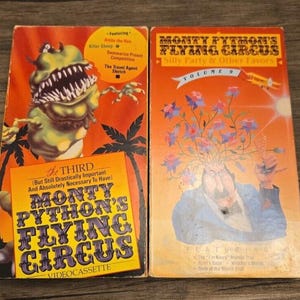 Pode incluir: Duas cassetes de vídeo Monty Python's Flying Circus com capas cor de laranja. A cassete da esquerda apresenta um monstro verde e texto incluindo "The Third" e "Monty Python's Flying Circus". A da direita mostra um homem com flores e "Volume 9".