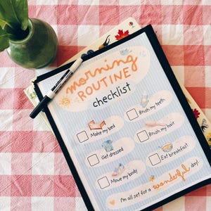 May include: A morning routine checklist in a black frame with a white dry-erase marker. The light blue chart features orange text and illustrations for tasks including brush my teeth, make my bed, brush my hair, get dressed, eat breakfast, and move my body.