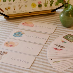 Op de afbeelding: Een verzameling educatieve kaarten met aquarel illustraties en tekstlabels, waaronder "Rest Time", "Unit Study", "Art" en "Cooking". Een botanische kaart en een groene vaas staan op de achtergrond. De kaarten liggen op een gestreept oppervlak.