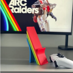 May include: A vibrant, multi-coloured phone stand with a red top, yellow, green, and blue layers. A white game controller is on the right. The screen in the background displays the text "ARC Raiders".