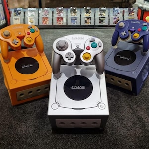 May include: Three Nintendo GameCube consoles in orange, silver, and purple. Each console has a matching controller. The consoles are displayed on a dark grey surface, with a collection of game cartridges in the background.