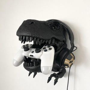 May include: A black dinosaur head wall mount with a white game controller in its mouth. Black headphones with gold detailing are on the side. The dinosaur has sharp teeth and a textured surface.