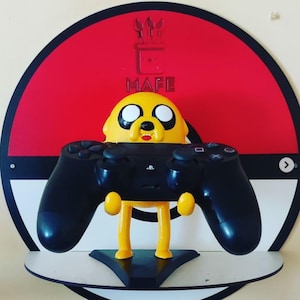 May include: A novelty gaming controller holder featuring a yellow cartoon character holding a black PlayStation controller. The holder is set against a red and white circular background. The shelf is white.