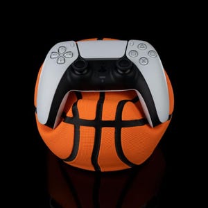 May include: A white and black PlayStation 5 controller sits on an orange basketball. The controller has a modern design with various buttons and directional pads. The basketball features black lines, creating a classic sports ball appearance.